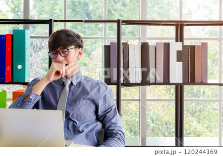 Businessmanl in glasses sitting at office desk and yawning while working with laptop. Bookshelf with colorful binders and books on background Businessmanl in glasses sitting at office desk and yawning while working with laptop. Bookshelf with colorful binders and books on background 120244169