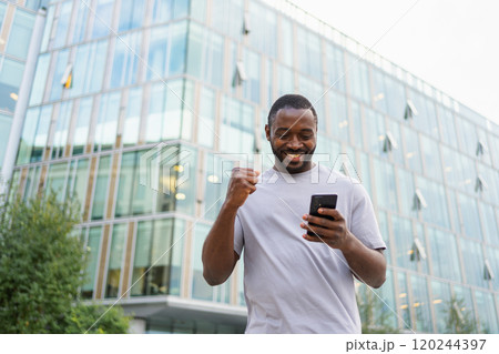 Happy African American man euphoric winner with smartphone on street in city. Person guy looking at cell phone reading great news getting good result winning online bid feeling amazed. Winning gesture 120244397