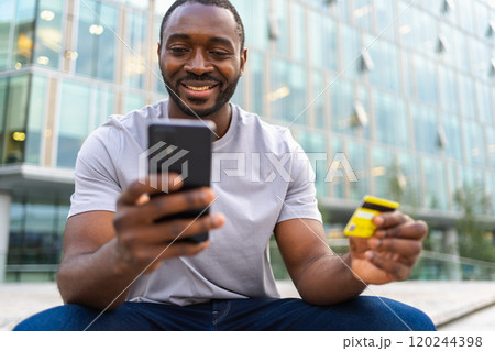 African American man shopping online holding smartphone paying with gold credit card Guy on urban street in city buying on Internet enter credit card details Online shopping ecommerce delivery service 120244398