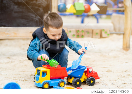 Little cute toddler boy three years old plays in the sandbox on a spring day. Outdoor development activities for kids. Toy cars and shovel 120244509