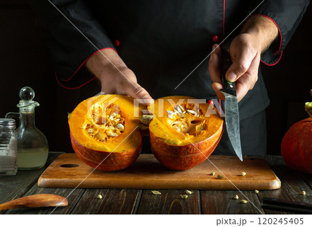 A skilled chef slices a ripe pumpkin in half, revealing its vibrant orange flesh and seeds. The rustic kitchen setting enhances the charm of this seasonal preparation A skilled chef slices a ripe pumpkin in half, revealing its vibrant orange flesh and seeds. The rustic kitchen setting enhances the charm of this seasonal preparation 120245405