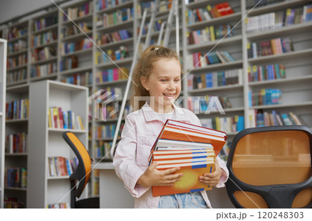 Cheerful smiling girl kid with stack of book standing over library background 120248303