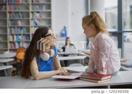 Young woman tutor teaching little girl to read in public library 120248308