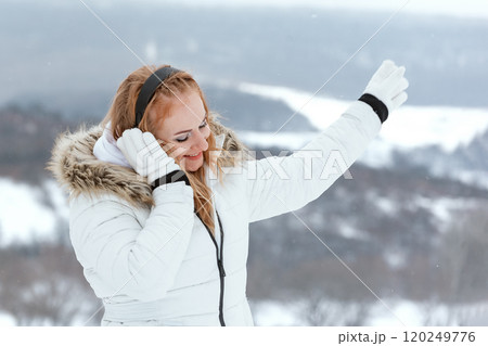 Music lover listens records on snowy winter day outside. Red-haired young woman wearing headphones in mountains in winter. Music lover listens records on snowy winter day outside. Red-haired young woman wearing headphones in mountains in winter. 120249776