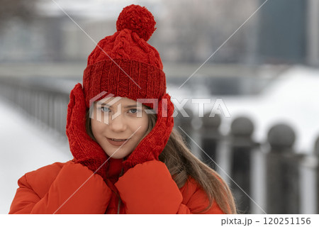 Cute girl in red jacket and set of knitted hats and mittens poses in winter outside. Trendy winter look for every day 120251156