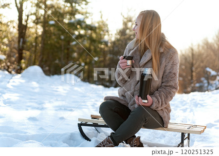 Young ginger woman drinks hot tea from thermos cup in snowy winter forest. Warm up in frozen winter woods. 120251325