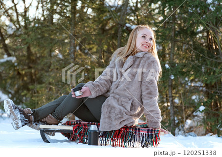 Positive young woman sitting on sled on pine trees background. Outdoor activities. Winter time. 120251338
