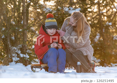 Young woman with son in winter forest on picnic drink hot tea in warm light. Happy family spends weekends outside the city. 120251495