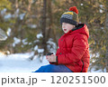 Boy in red jacket and hat sitting on the snow with a cup in his hand, sits and looks into the distance 120251500