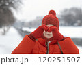 Portrait of a beautiful girl in a red jacket and hat shows heart sign with her palms on a background of a winter park 120251507