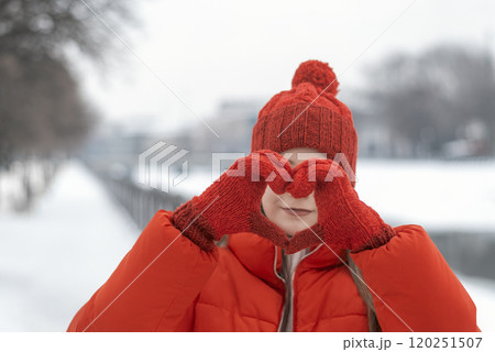 Portrait of a beautiful girl in a red jacket and hat shows heart sign with her palms on a background of a winter park 120251507