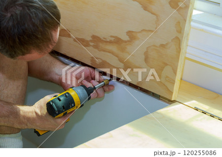 Worker is focused on securing plywood panels hinges to surface using power drill. 120255086