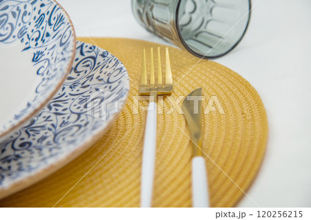 A white soup plate with a blue pattern on the edges stands on a blue-and-white patterned serving plate in close-up on a yellow straw napkin, next to it are cutlery, a knife and fork, behind them is a A white soup plate with a blue pattern on the edges stands on a blue-and-white patterned serving plate in close-up on a yellow straw napkin, next to it are cutlery, a knife and fork, behind them is a 120256215