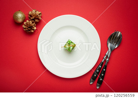 Small green christmas gift on white plate with golden pine cones and christmas ball ornament on red background Small green christmas gift on white plate with golden pine cones and christmas ball ornament on red background 120256559