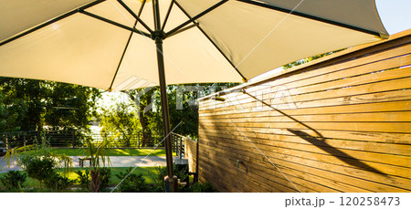 Sunlit outdoor umbrella and wooden fence with natural greenery. Concept of shade, privacy, and peaceful outdoor retreat 120258473
