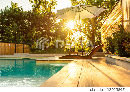 Sunlit poolside with wooden deck and lounge chair under umbrella. Concept of serene outdoor relaxation and luxury retreat 120258474