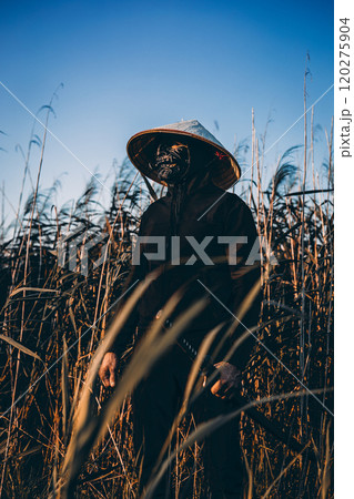 A samurai wearing a mask, conical hat and black clothes stands in the tall grass holding a katana, creating a dramatic and mysterious scene. Samurai with a katana ready to repel any attack. 120275904