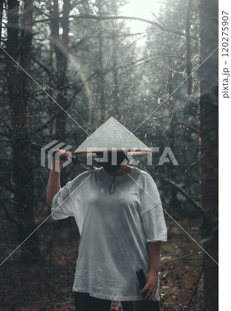 Samurai with katana in forest during rain in sun rays. Mysterious man in black mask and asian conical hat standing in forest holding katana sword in light rain. Samurai with katana in forest during rain in sun rays. Mysterious man in black mask and asian conical hat standing in forest holding katana sword in light rain. 120275907