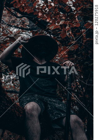 A masked cosplayer sits on a tree branch in the forest, holding a katana and adjusting his hat. A samurai with a katana takes a break from posing for photos. A warrior with a katana in the forest. 120275918