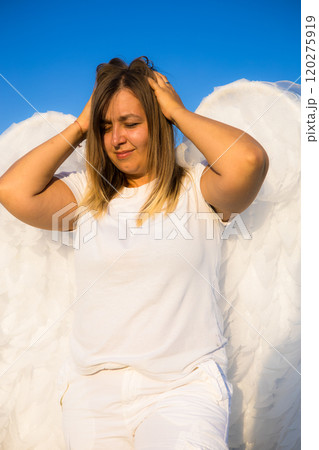 An angel in despair holds her head from what is happening in the world. A blonde in white clothes and with big white angel wings touches her hair with both hands under a clear blue sky. An angel in despair holds her head from what is happening in the world. A blonde in white clothes and with big white angel wings touches her hair with both hands under a clear blue sky. 120275919