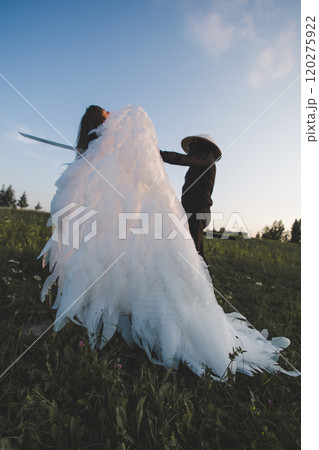 Samurai stabs a beautiful white angel with a katana. A samurai with a katana attacks an angel with large white wings on a green field at sunset, creating a dramatic and surreal scene. 120275922