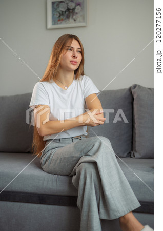 Young woman seated on a gray couch showing signs of discomfort while wearing a casual white t-shirt and gray pants in a cozy indoor setting 120277156