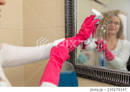 A woman is diligently cleaning a mirror using gloves and a spray cleaner for a pristine shine A woman is diligently cleaning a mirror using gloves and a spray cleaner for a pristine shine 120279147