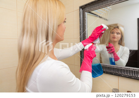 A woman is thoroughly cleaning a mirror using a spray bottle in her bright bathroom setting 120279153