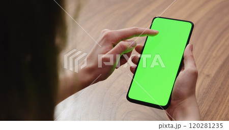 Young woman sitting at a table and using a smartphone with vertical green screen 120281235