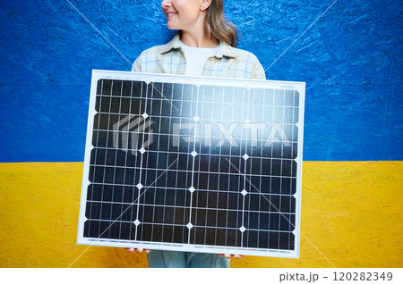 Woman holding solar panel for charging. Smiling female wears plaid shirt over white top, standing against vibrant blue and yellow Ukrainian flag background, symbolizing sense of pride and energy. Woman holding solar panel for charging. Smiling female wears plaid shirt over white top, standing against vibrant blue and yellow Ukrainian flag background, symbolizing sense of pride and energy. 120282349