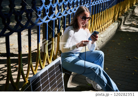 Happy woman connecting smartphone to photovoltaic solar panel. Integration of sustainable renewable energy into everyday life, demonstrating practical use of solar power for charging devices. 120282351