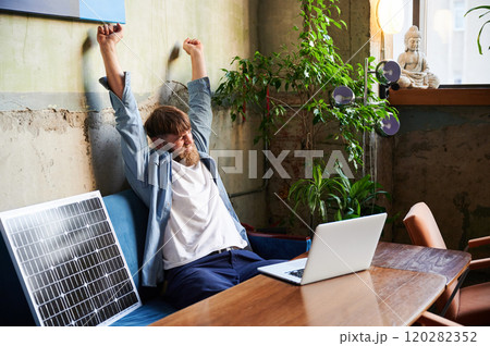 Bearded man stretches with arms overhead while working on laptop at wooden table. Solar panel nearby, emphasizing eco-friendly workspace that combines technology and sustainability in cozy environment 120282352