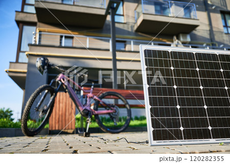 Pink electric mountain bike parked next to wooden bench, connected to solar panel for charging on a blurred background. Bike rests on paved area surrounded by greenery and grass. 120282355
