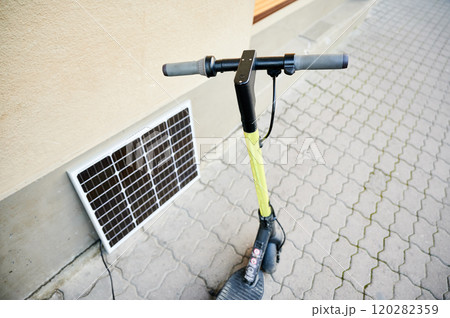 Electric scooter with yellow handlebar parked on patterned sidewalk next to beige wall. Solar panel leans against wall, connected to scooter, highlighting clean energy charging setup in urban setting. Electric scooter with yellow handlebar parked on patterned sidewalk next to beige wall. Solar panel leans against wall, connected to scooter, highlighting clean energy charging setup in urban setting. 120282359