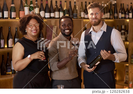 Portrait of multiracial company of professional sommeliers during degustation red wine in wine cellar fully equipped with wooden racks and cabins for wine bottles 120282638