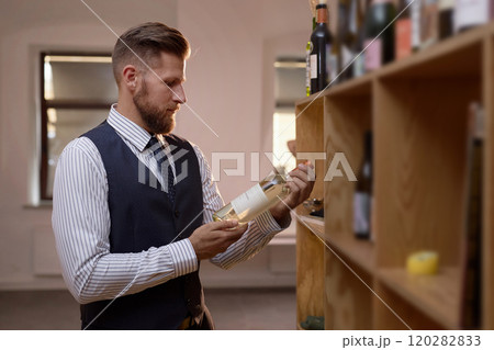 Professional Caucasian sommelier wearing smart clothes standing next to wooden cabins full of bottles while reading wine bottle label 120282833