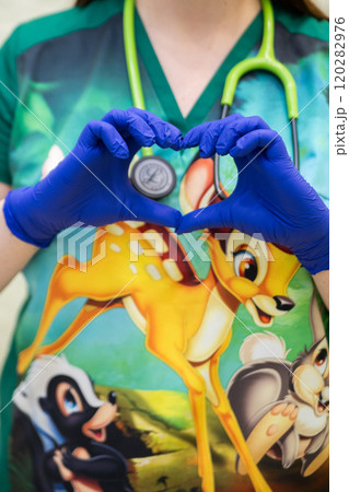 A healthcare professional wearing blue gloves forms a heart shape with their hands over a shirt featuring a colorful cartoon character, with a green stethoscope draped around their neck. 120282976