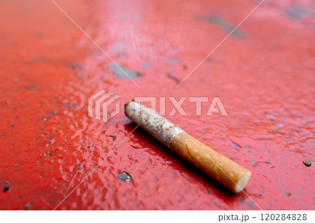 Close-up of a discarded cigarette lying on a wet red ground surface with visible water droplets Close-up of a discarded cigarette lying on a wet red ground surface with visible water droplets 120284828
