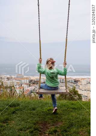 A young Caucasian girl in a green jacket on a swing overlooking a coastal town and ocean, filmed from the back with no face visible A young Caucasian girl in a green jacket on a swing overlooking a coastal town and ocean, filmed from the back with no face visible 120284837