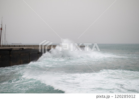 Rough ocean waves crashing powerfully against a concrete pier on a foggy day, with water spraying over the edge and creating a dramatic scene 120285012