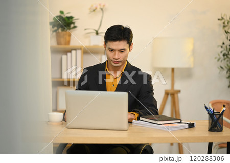 Concentrated asian businessman in black suit working on laptop at office Concentrated asian businessman in black suit working on laptop at office 120288306