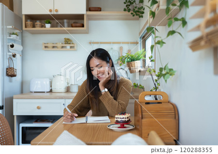 Happy young adult woman enjoying a peaceful moment with journaling in kitchen 120288355