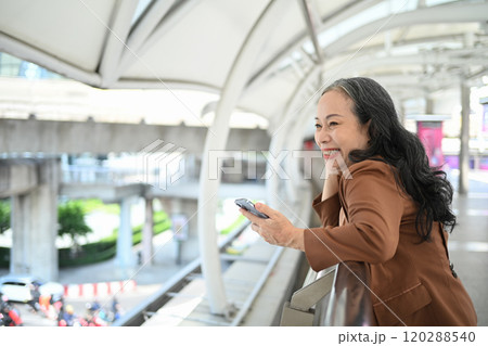 Happy mature businesswoman holding smartphone standing in a covered outdoor walkway 120288540