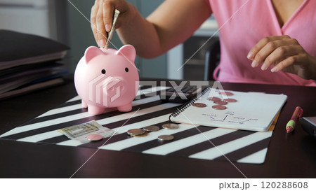A young woman using a calculator to count coins and banknotes while sitting at a kitchen table, placing money into a pink piggy bank. Savings and budgeting concept A young woman using a calculator to count coins and banknotes while sitting at a kitchen table, placing money into a pink piggy bank. Savings and budgeting concept 120288608