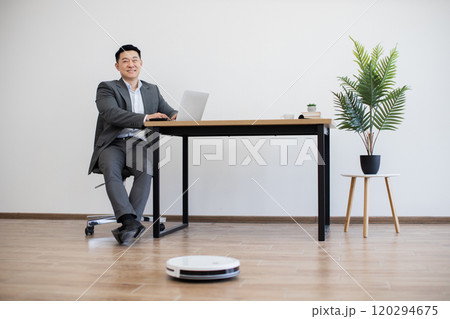Asian businessman working on laptop with robot vacuum cleaning floor Asian businessman working on laptop with robot vacuum cleaning floor 120294675