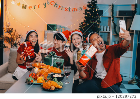 Group of young Asian man and women as friends having fun at a New Year's celebration, holding gift boxes standing by Christmas tree decoration, midnight countdown Party at home with holiday season. 120297272