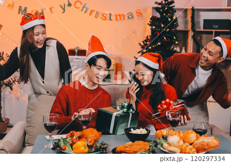 Group of young Asian man and women as friends having fun at a New Year's celebration, holding gift boxes standing by Christmas tree decoration, midnight countdown Party at home with holiday season. 120297334