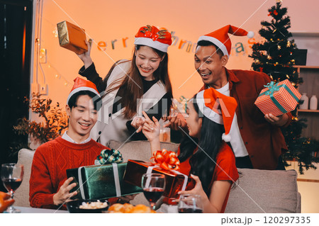Group of young Asian man and women as friends having fun at a New Year's celebration, holding gift boxes standing by Christmas tree decoration, midnight countdown Party at home with holiday season. 120297335