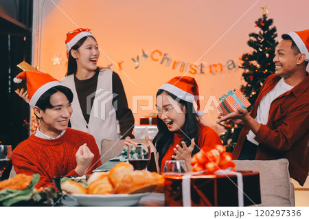 Group of young Asian man and women as friends having fun at a New Year's celebration, holding gift boxes standing by Christmas tree decoration, midnight countdown Party at home with holiday season. 120297336