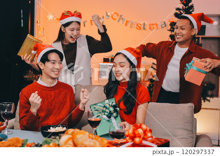 Group of young Asian man and women as friends having fun at a New Year's celebration, holding gift boxes standing by Christmas tree decoration, midnight countdown Party at home with holiday season. 120297337